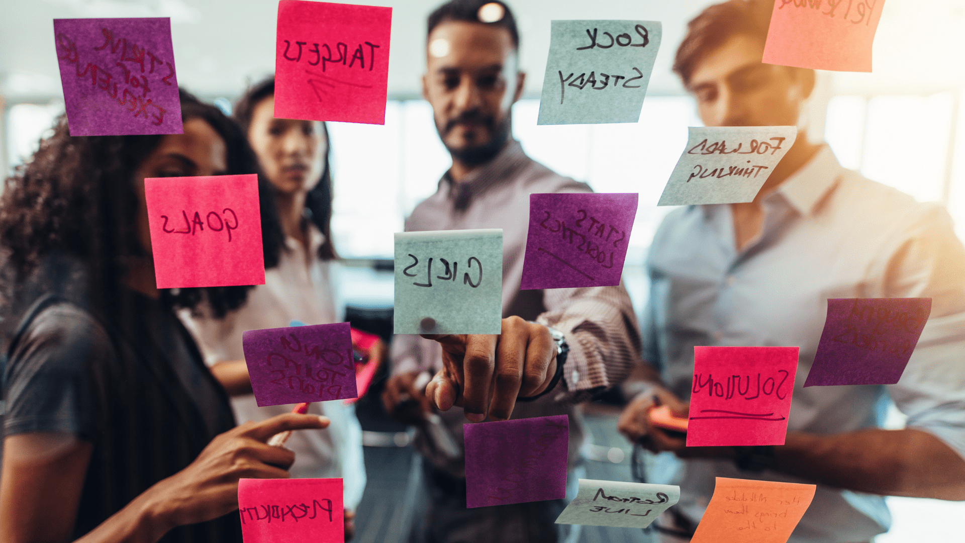 A group of 4 people brainstorming together , pointing at sticky notes on a glass wall.