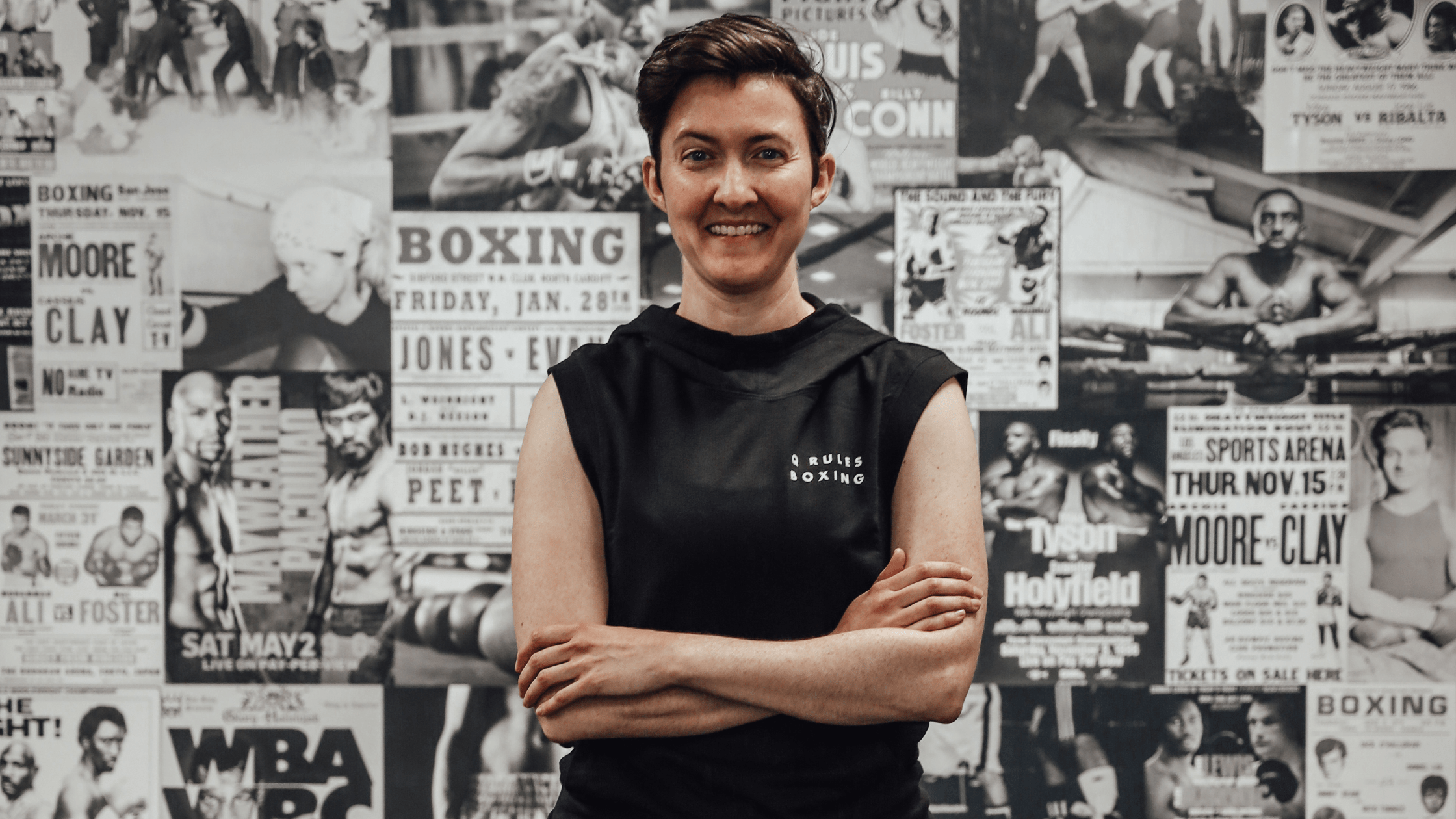 A photo of Lisa Buchanan standing arms crossed in front of a wall of black and white photos of boxers.